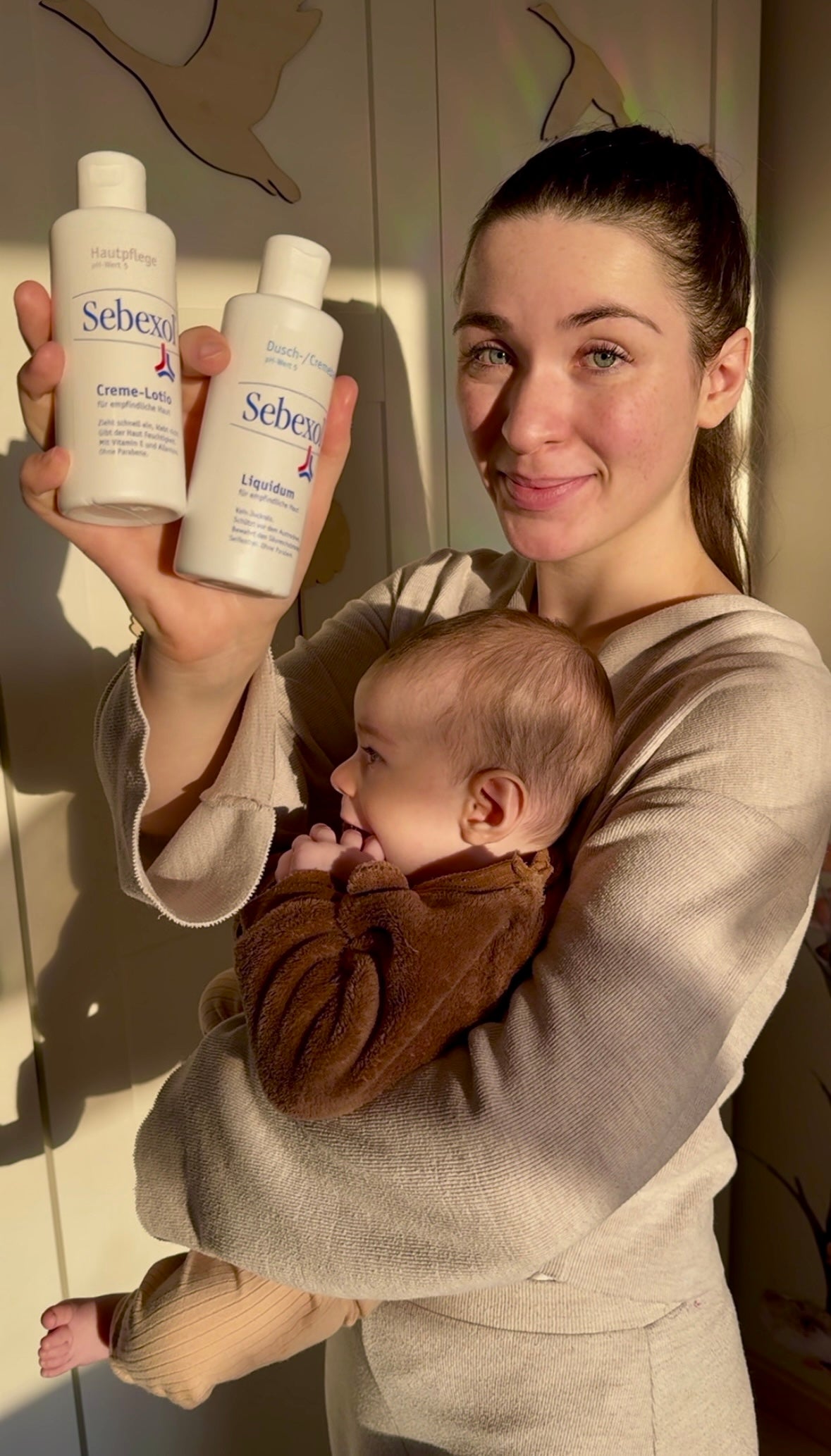 Junge Frau hält Sebexol Creme-Lotio und Sebexol Liquidum lächelnd in die Kamera, auf ihrem Arm ein Baby.